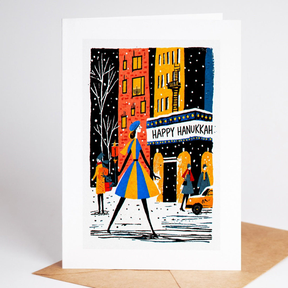 New York Hanukkah card standing with envelope, a woman in a blue and gold striped coat crossing under a Happy Hanukkah s