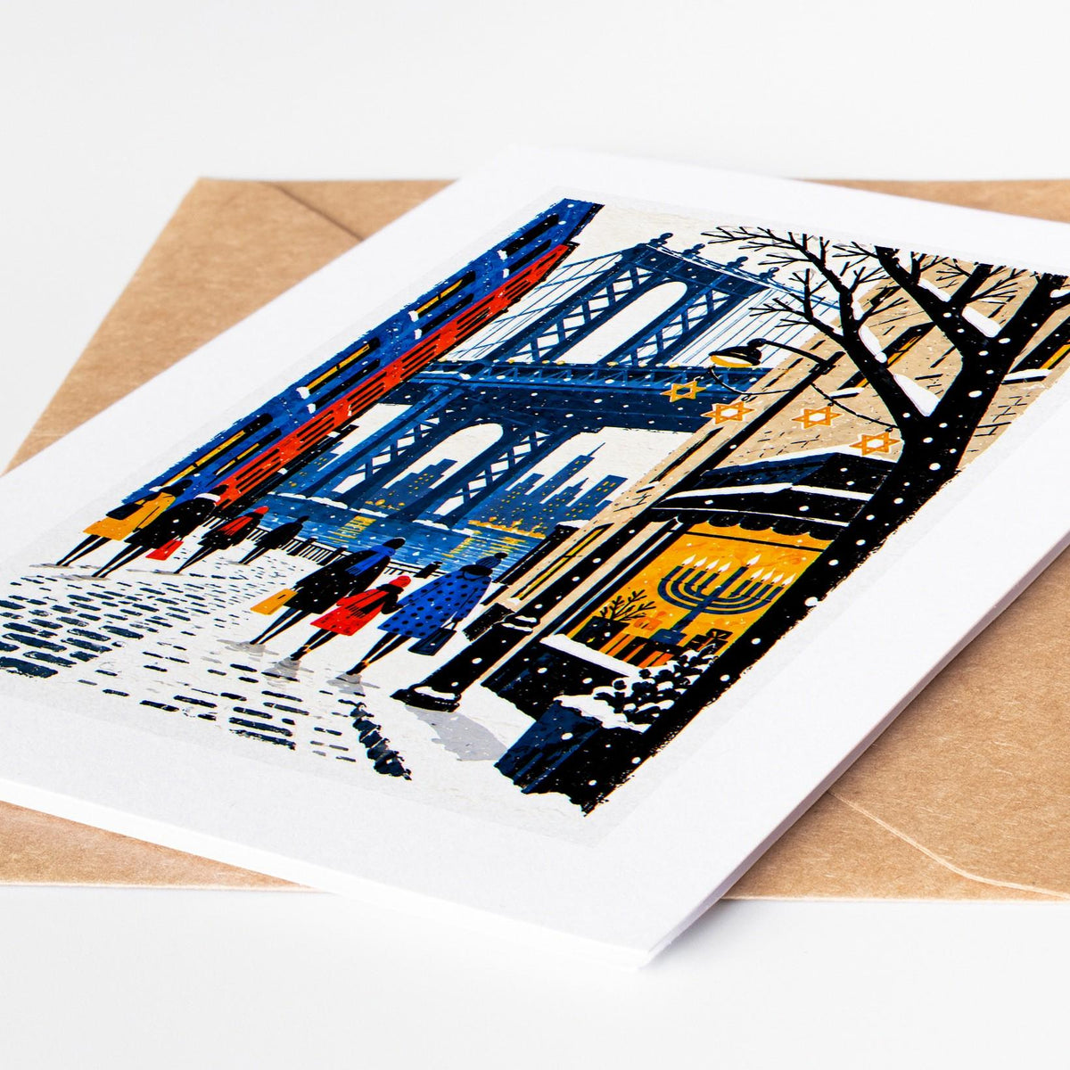 Brooklyn Hanukkah card lying on a kraft envelope, folk art DUMBO under the Manhattan Bridge with a lit menorah in a shop